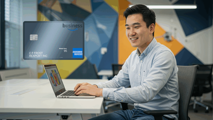 An Asian man in a light blue shirt is smiling while working on a laptop in a modern office with geometric wall designs. In the top left corner, there is a graphic of an Amazon Business American Express credit card