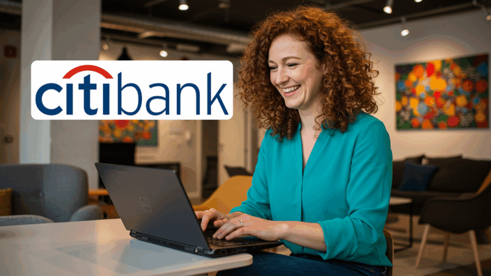 A smiling woman with curly red hair wearing a teal blouse is working on a laptop in a modern, colorful office space. The Citibank logo is displayed in the upper left corner.