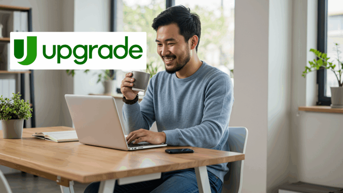 A smiling man in a gray sweater drinking coffee and working on a laptop at a wooden desk in a bright, modern room, with the Upgrade logo overlaid on the left side.