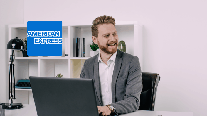 Bearded man in a gray suit sitting at a desk with a laptop, smiling and looking to the side in a bright office space. The American Express logo is displayed in the background.