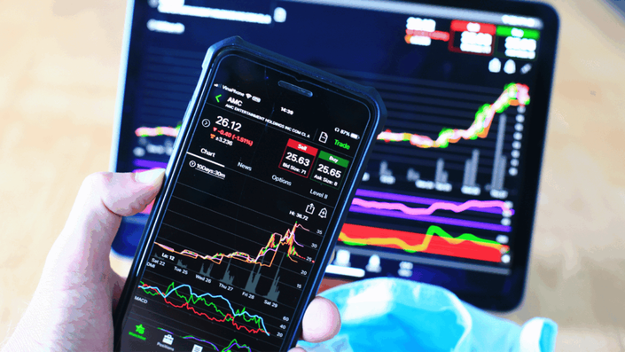 Hand holding a smartphone displaying a stock trading app with charts and figures, with a tablet showing more financial data in the background, representing an investor monitoring Stock Market Volatility and their portfolio.