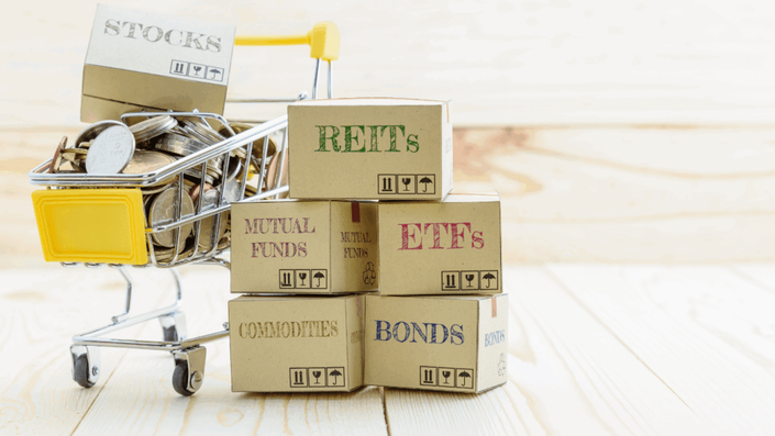 Miniature shopping cart with coins and a box labeled 'STOCKS' alongside stacked boxes labeled 'REITs', 'MUTUAL FUNDS', 'ETFs', 'COMMODITIES', and 'BONDS', symbolizing investment diversification as a strategy against Stock Market Volatility.